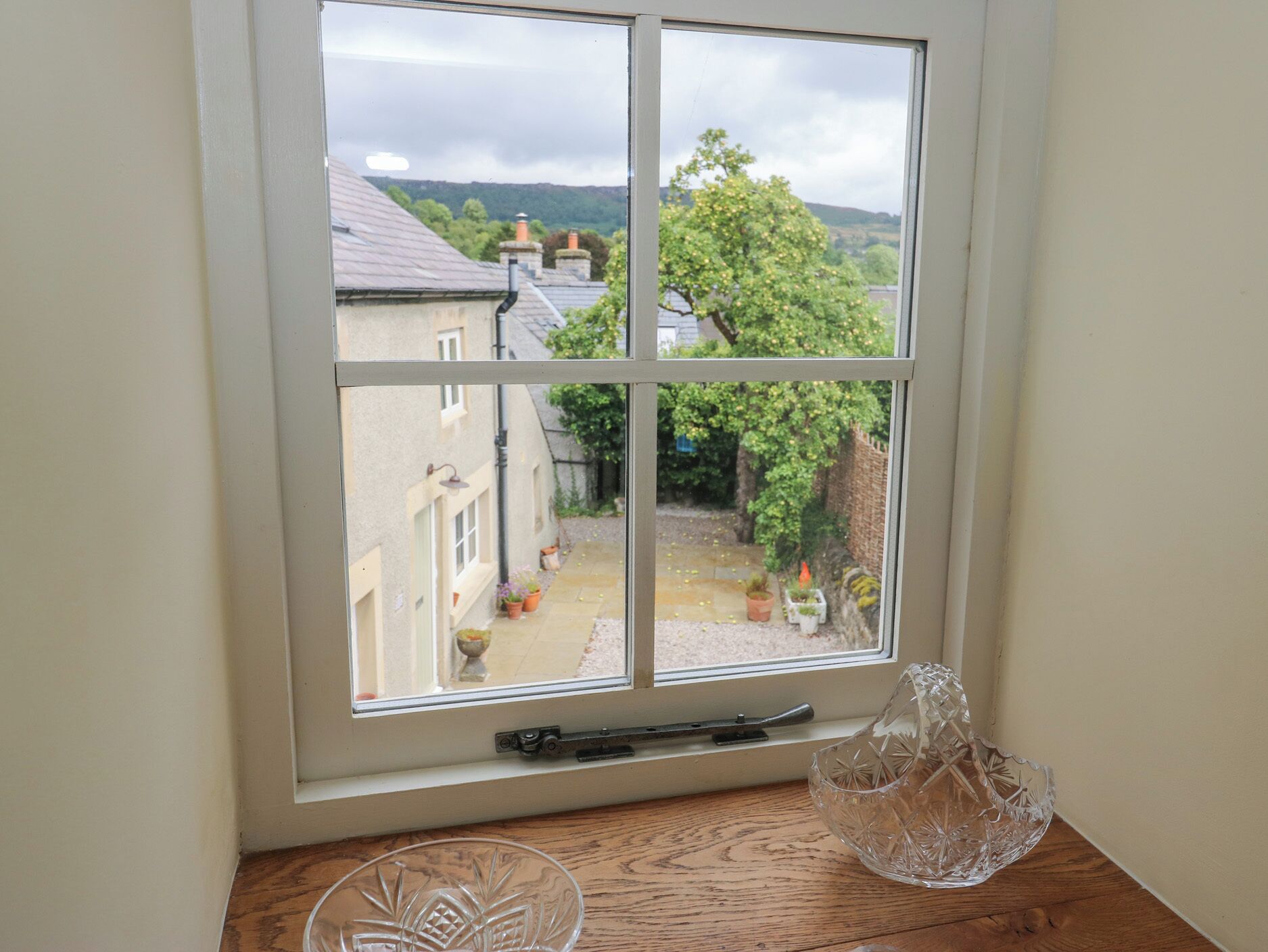 A window with a view of a garden and buildings, a wooden table with a glass bowl, and a window sill with a small decorative item.