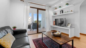 En moderne stue med en lædersofa, et sofabord og et fjernsyn på et hvidt skab. Der er en balkon med udsigt over bygninger og et gelænder.