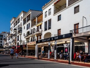 A street with outdoor seating at a café and bar, surrounded by multi-story buildings with balconies. - Sea Views Apartment with Terrace and Pools (Puerto de la Duquesa)