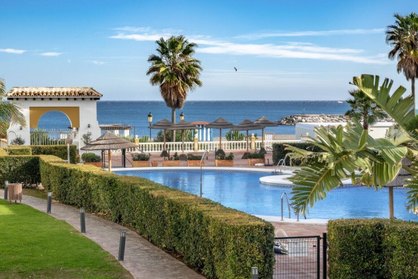 A pool area with a clear view of the sea, a palm tree, and a building with a tiled roof.