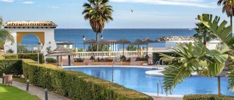 A pool area with a clear view of the sea, a palm tree, and a building with a tiled roof.