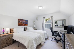 Un dormitorio con dos camas, un escritorio con computadora, una silla, una lámpara, un cuadro y una ventana con cortinas.