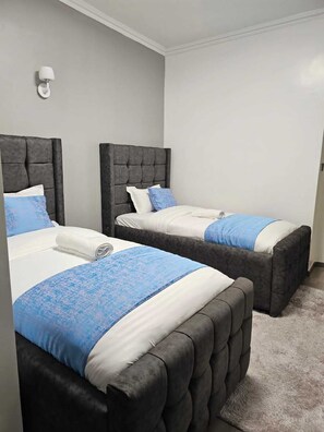 Quarto de hotel com duas camas, cada uma com cabeceira estofada, parede branca e uma pequena luminária fixada na parede.