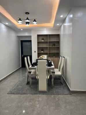 A modern dining area with a table set for four, surrounded by chairs, and a built-in shelving unit against the wall. - Cozy Gardens View Palace Apartment (Nairobi)
