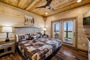 Ein Schlafzimmer mit Holzbalkendecke, einem Bett mit Quilt, zwei Nachttischlampen, einem Bild an der Wand und einem Fenster mit Jalousien.