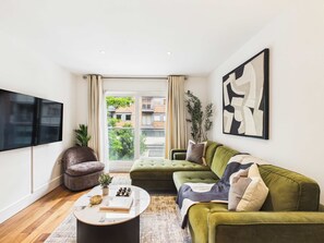 Ein modernes Wohnzimmer mit einem Flachbildfernseher, einem grünen Sofa, einem runden Couchtisch und einem großen Fenster mit Blick auf Gebäude und Grünflächen.