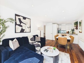 A modern living room with a dark blue sofa, a round coffee table, and a dining area with a white table and yellow chairs. - Comfy 2 Bedroom Westminster Apartment (Greater London)