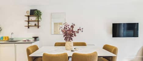 A dining area with a white table, four chairs, a vase with flowers, a mirror, and a TV on the wall.