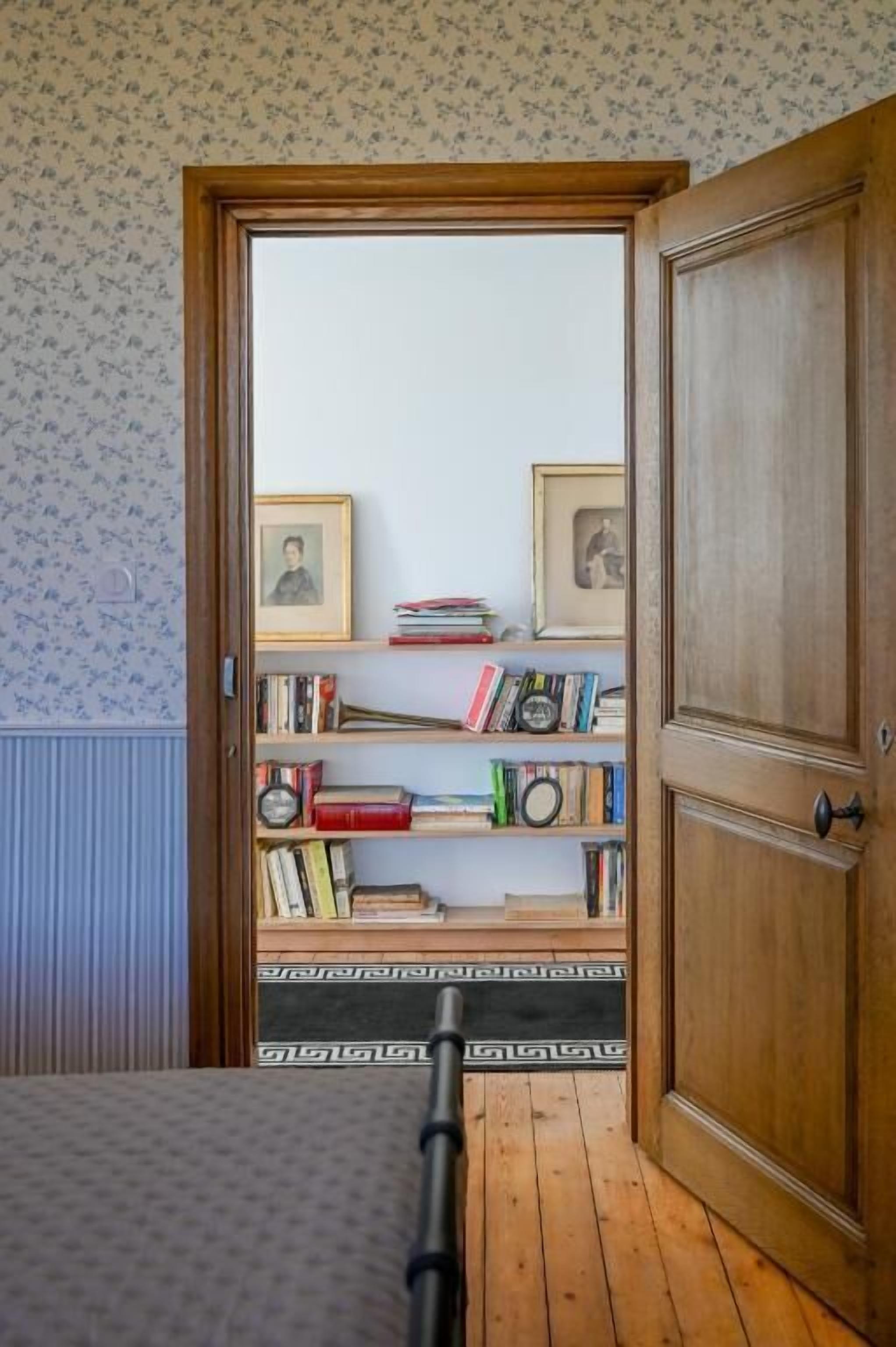A room with a wooden door, a bookshelf with books and framed pictures, and a patterned floor.