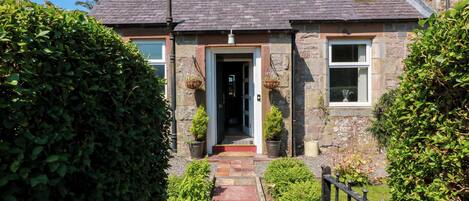A stone house with a tiled roof, a front door, and a small garden with a pathway leading to the entrance.