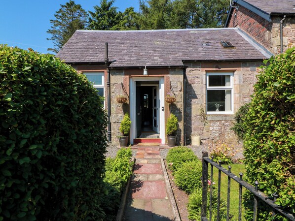 A stone house with a tiled roof, a front door, and a small garden with a pathway leading to the entrance.