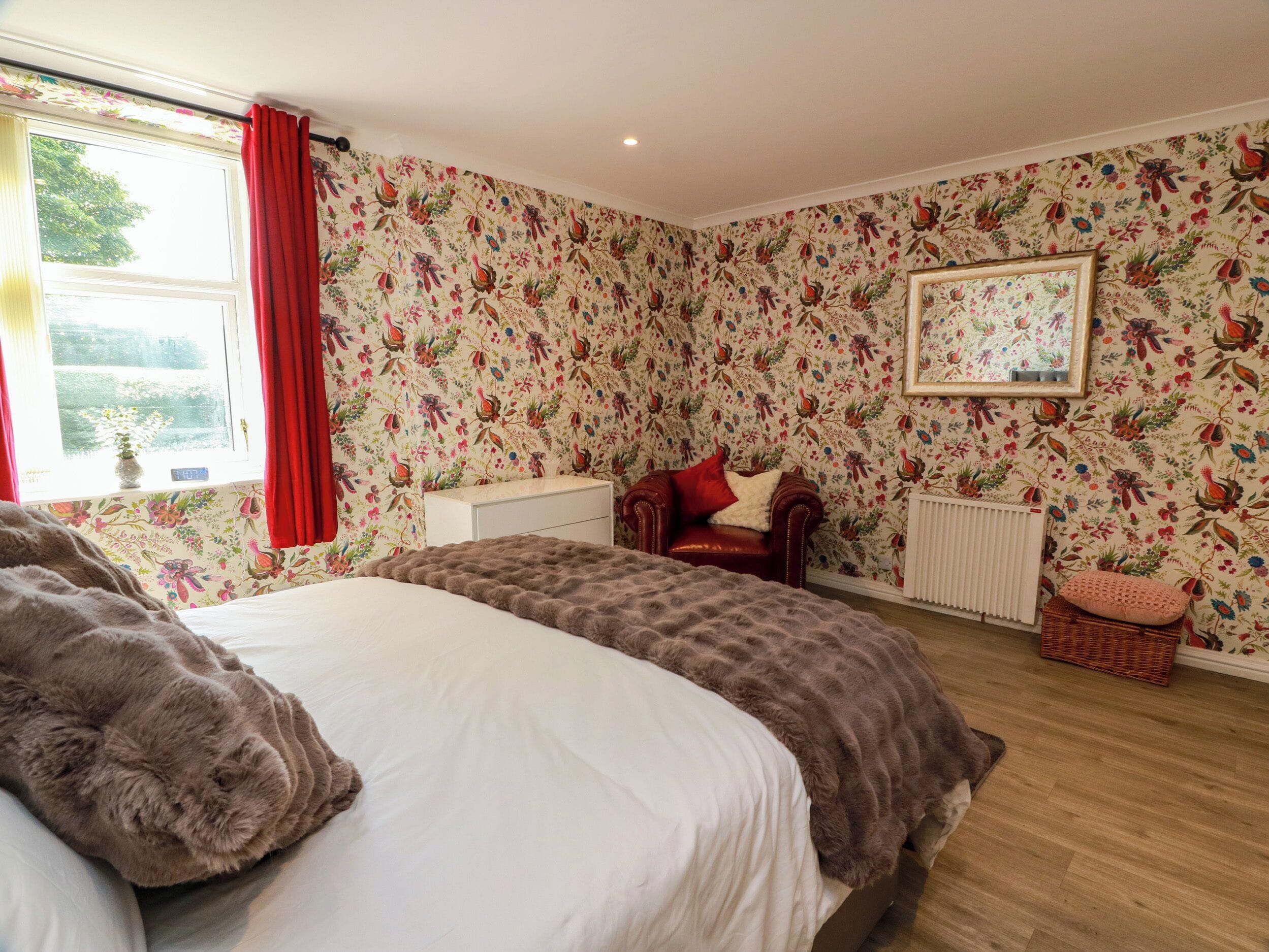 A bedroom with floral wallpaper, a bed with a brown blanket, a red chair, and a window with red curtains.