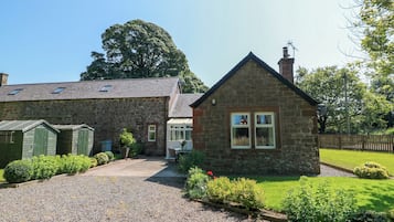 A stone house with a gravel driveway, a small garden, and a large tree in the background.