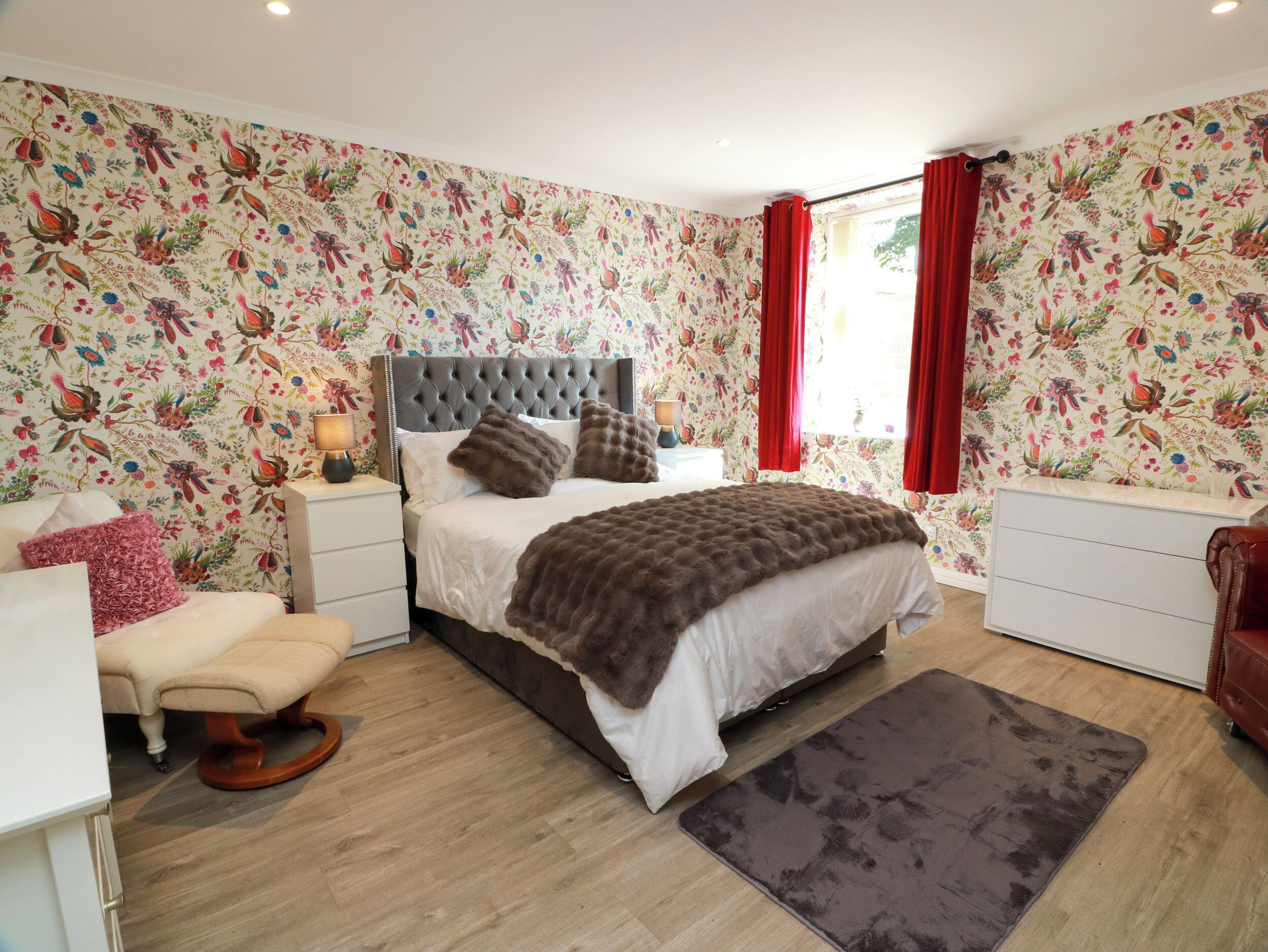 A bedroom with a tufted headboard, a bed with a brown blanket, a bedside table with a lamp, a chair, and a dresser.