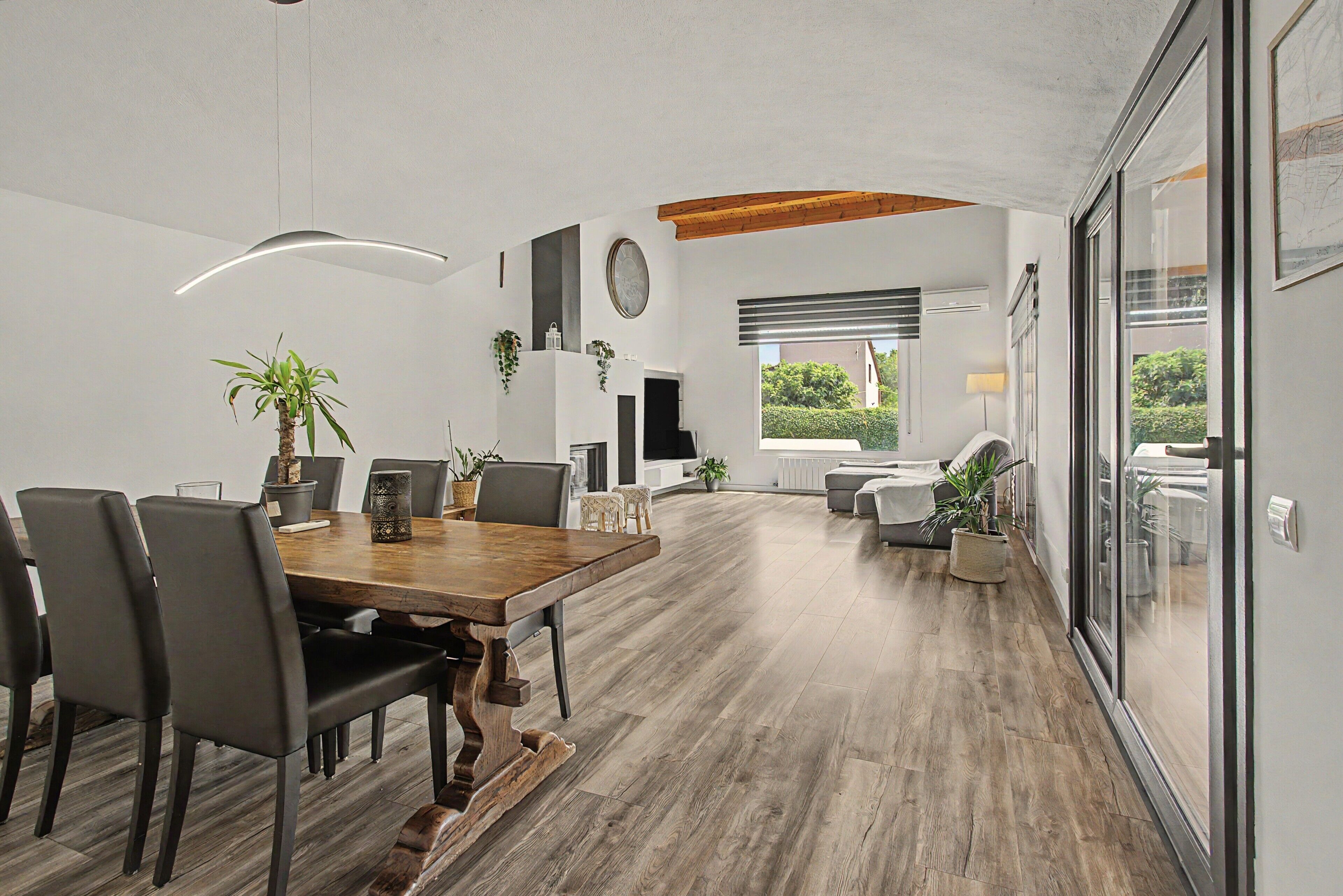Un vaste espace de salle à manger avec une table et des chaises en bois, une cuisine moderne en arrière-plan et une vue sur l’extérieur grâce à de grandes fenêtres.
