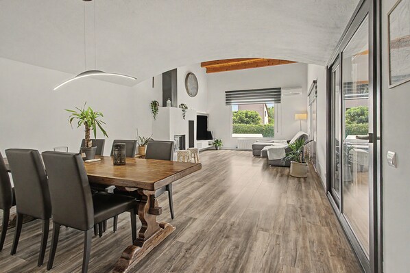 A spacious dining area with a wooden table and chairs, a modern kitchen in the background, and a view of the outdoors through large windows.