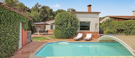 Una piscina con due lettini, un patio con tavolo e sedie, e un giardino con siepe e albero.