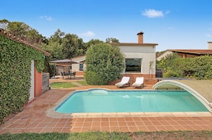 A swimming pool with two lounge chairs, a patio area with a table and chairs, and a garden with a hedge and a tree. - Cal Piano Vacation Home with Private Pool, Wi-Fi, and Air Conditioning (Palol de Revardit)