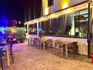An outdoor seating area with tables and chairs, a stone floor, and a large window overlooking the street.