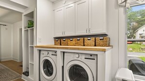 A laundry room with two front-loading washing machines, white cabinets above, and wicker baskets on a wooden shelf.