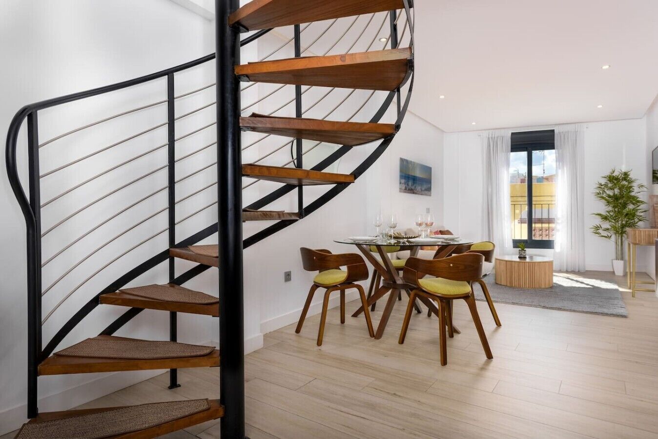 Un intérieur moderne avec un escalier en colimaçon, un coin repas avec des chaises en bois et une grande fenêtre laissant entrer la lumière naturelle.