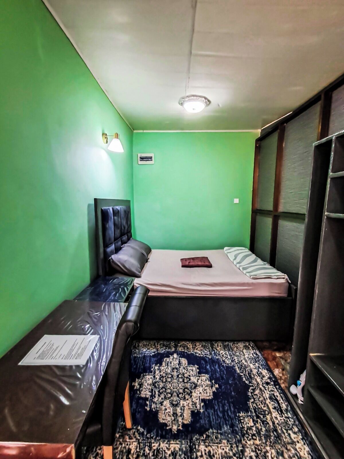 A small room with two beds, a green wall, a ceiling light, and a window.