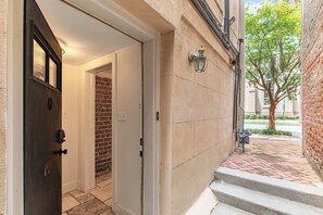 L’entrée d’un immeuble se caractérise par une porte sombre, un mur de briques et un trottoir pavé qui mène à une rue bordée d’arbres.
