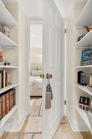 Een hal met witte muren en een deur die naar een slaapkamer leidt. Aan beide kanten staan planken met boeken en decoratieve objecten.