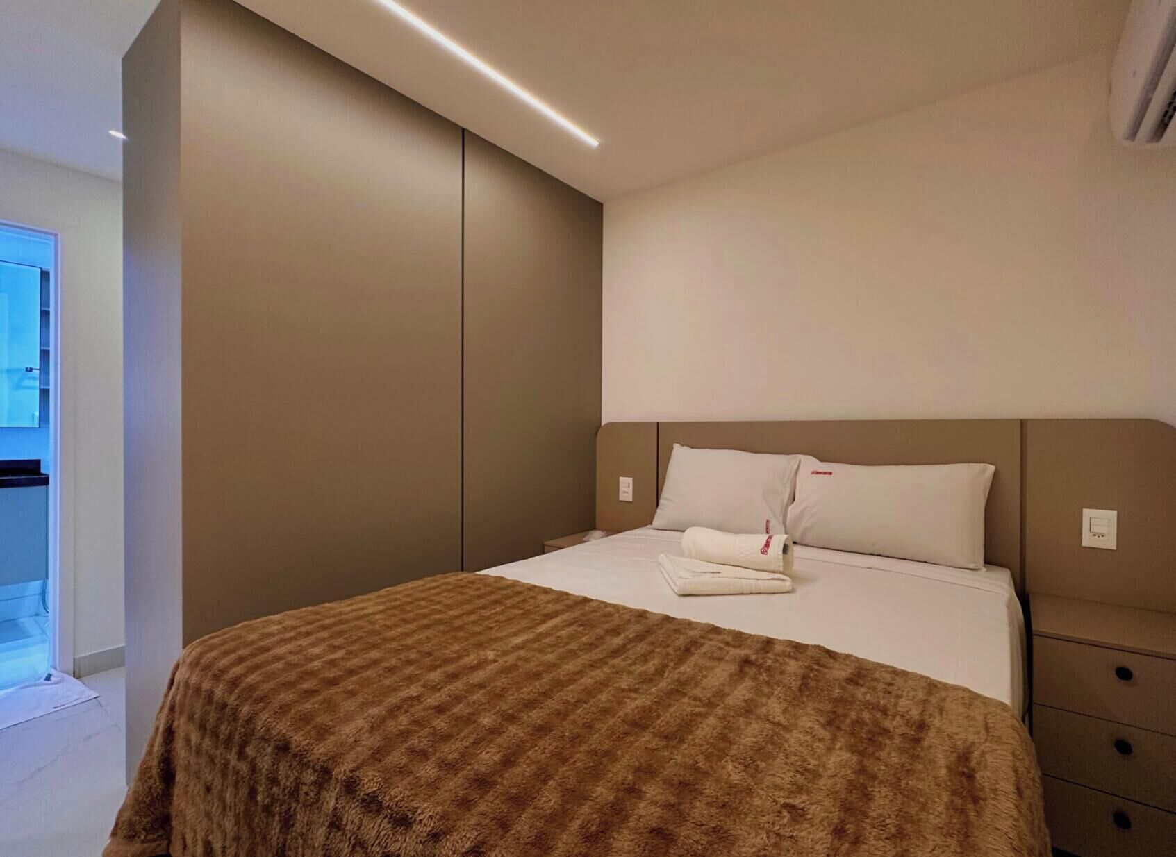 A modern hotel room with a large bed, a brown textured blanket, and a view of the outdoors.