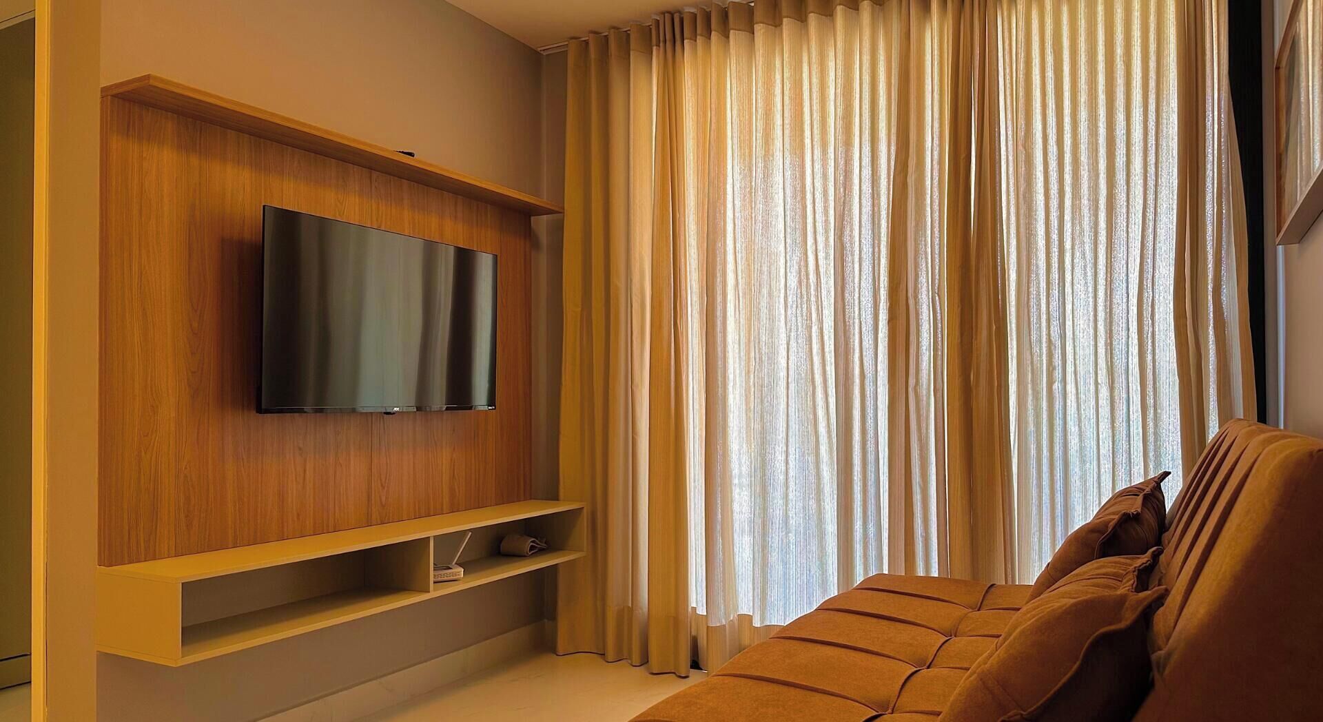 Uma sala de estar moderna com uma televisão de tela plana montada em uma parede de madeira, um sofá e uma janela com cortinas leves.