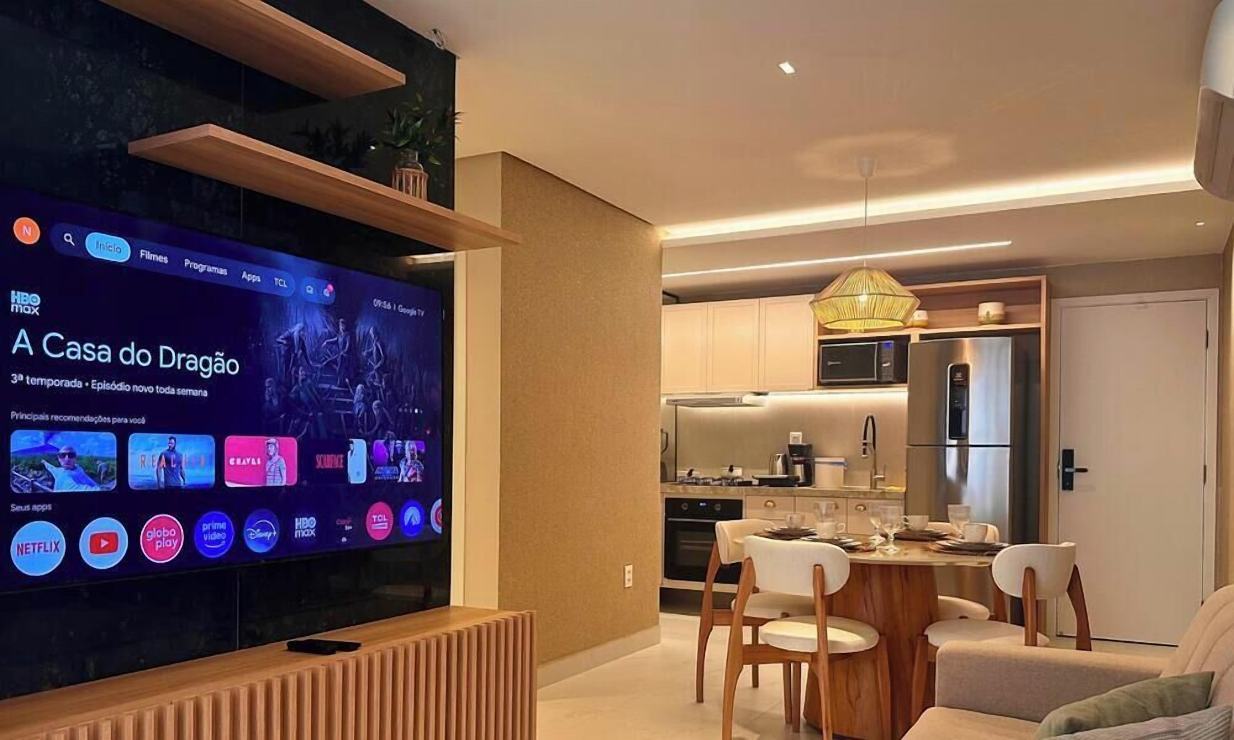 Una sala de estar moderna con un televisor de pantalla plana que muestra la interfaz de un servicio de streaming. Se observa un comedor con una mesa y sillas al fondo.