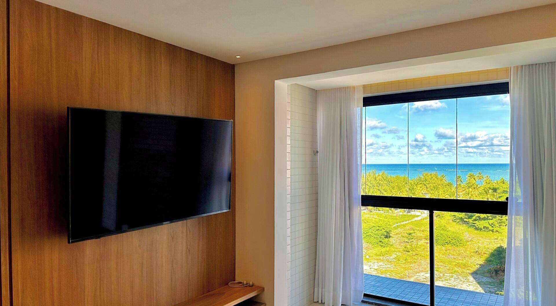 Ein modernes Zimmer mit einem an einer Holzwand befestigten Flachbildfernseher, einer Schiebeglastür, die auf einen Balkon mit Blick auf das Meer und Palmen führt.