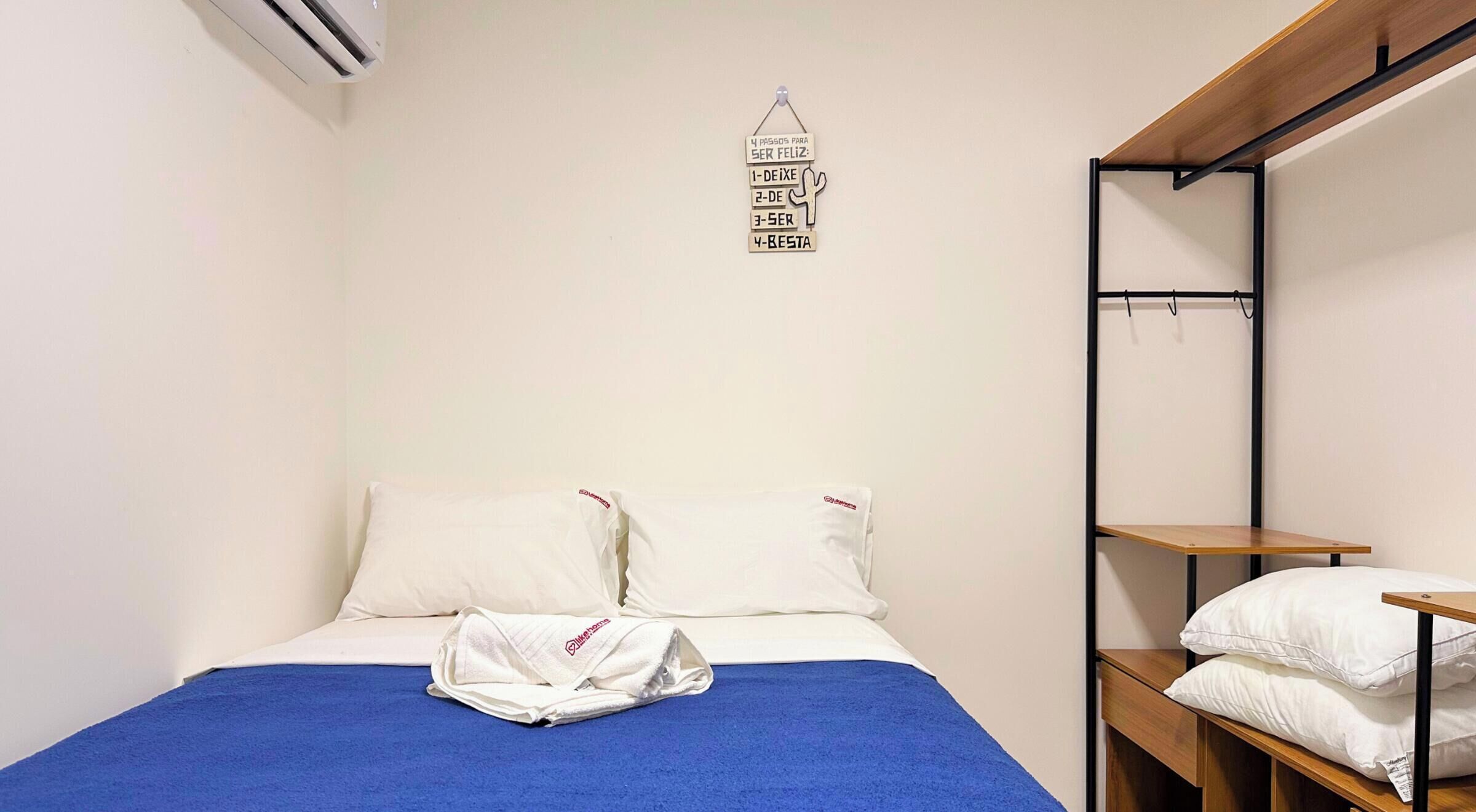Una cama individual con una colcha azul, dos almohadas blancas y una toalla blanca sobre la cama. Arriba de la cama, se observa un equipo de aire acondicionado instalado en la pared.