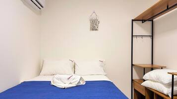 Una cama individual con una colcha azul, dos almohadas blancas y una toalla blanca sobre la cama. Arriba de la cama, se observa un equipo de aire acondicionado instalado en la pared.