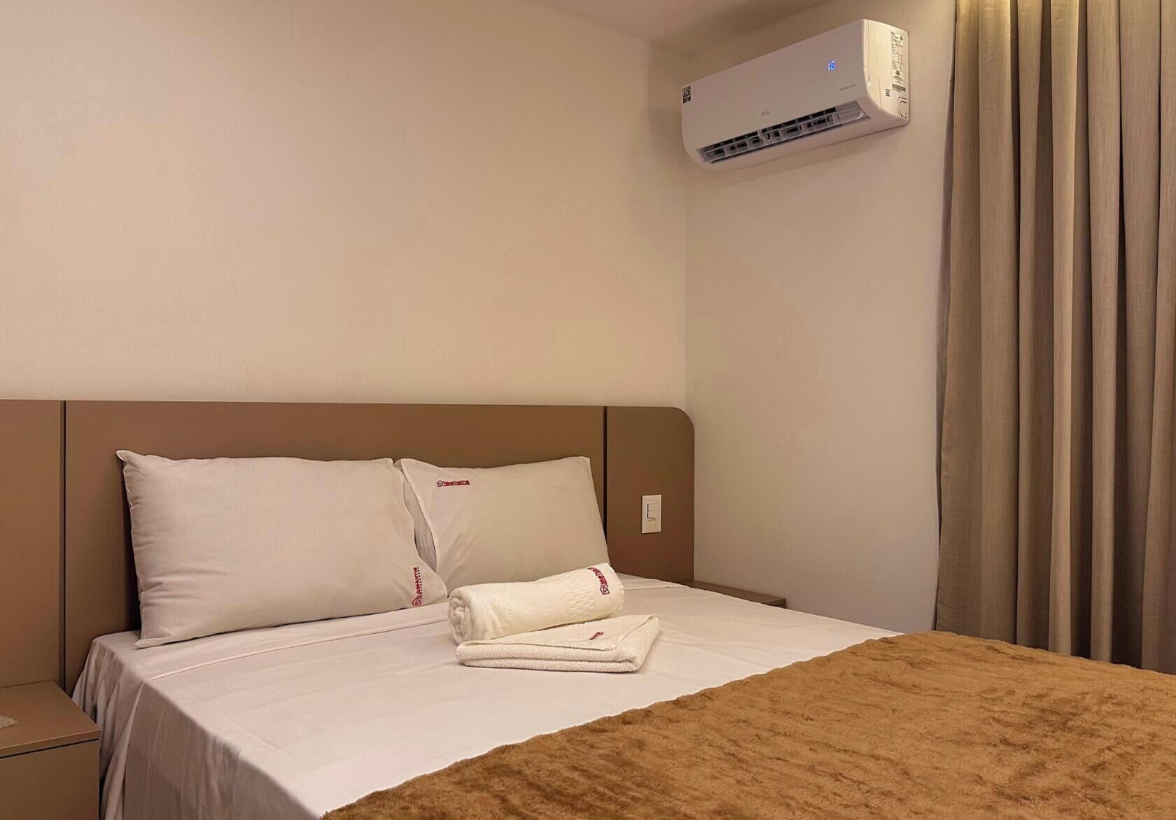 Habitación de hotel con cama, almohadas, una toalla doblada, equipo de aire acondicionado y una cortina.