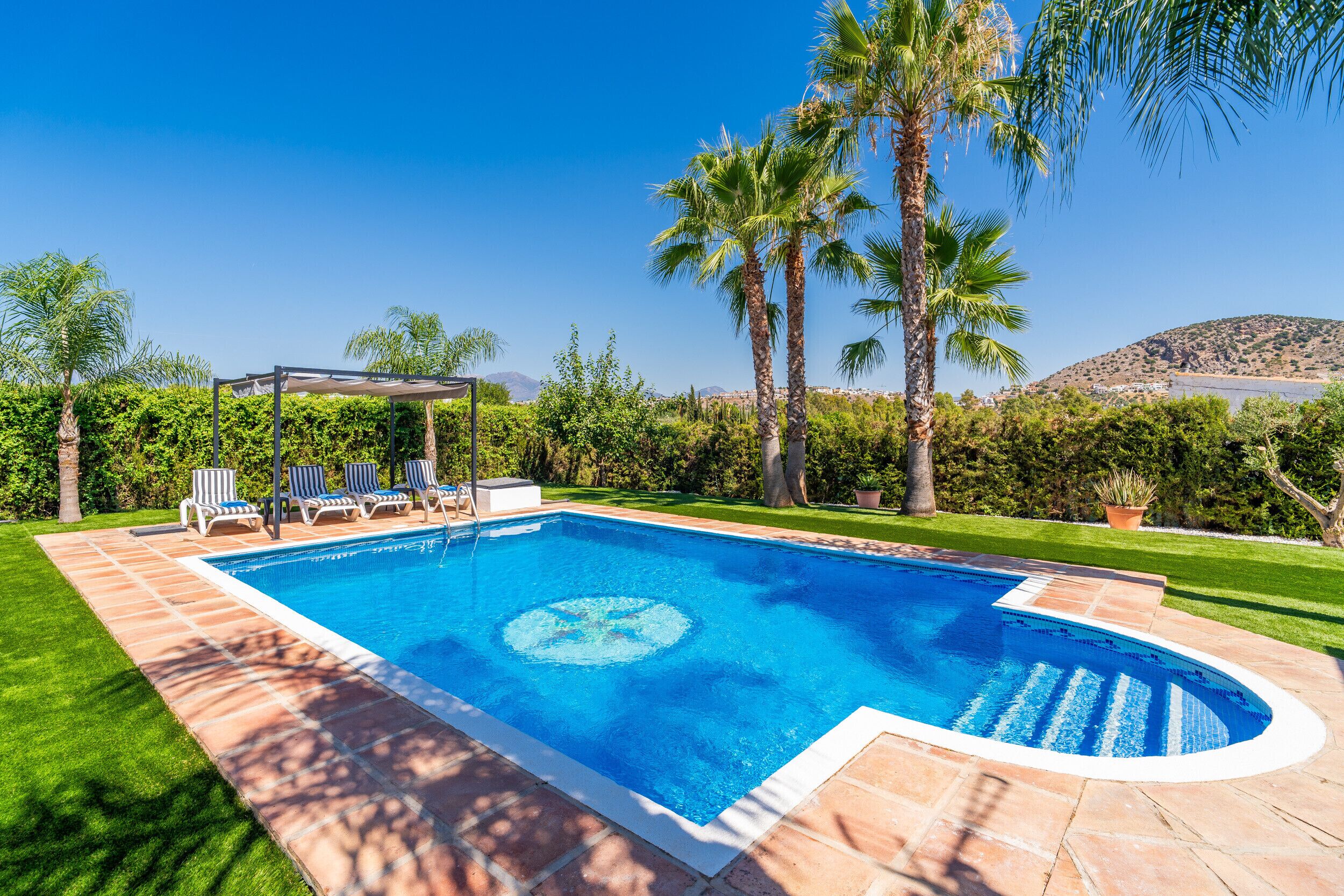 Ein rechteckiger Swimmingpool mit klar blauem Wasser, umgeben von einer gefliesten Liegefläche. Liegestühle und eine Pergola mit Dach sind vorhanden.