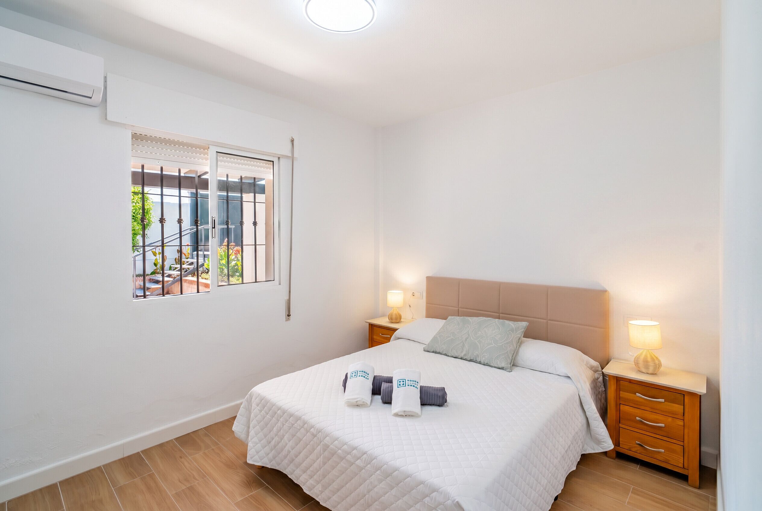 A hotel room with a bed, two bedside tables with lamps, a window with a view of the outside, and an air conditioning unit on the wall.