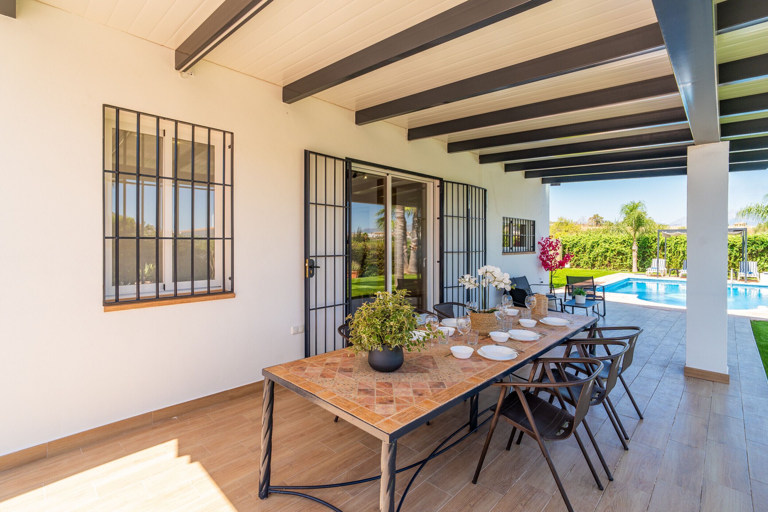 A covered outdoor dining area with a table set for four, surrounded by chairs, and a view of a swimming pool and greenery.