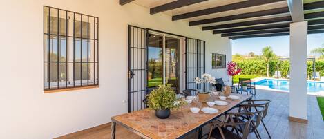 Un área de comedor al aire libre techada, con una mesa dispuesta para cuatro personas, sillas alrededor y vistas a una piscina y follaje.