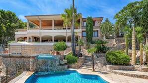 A swimming pool with a stone wall and palm trees, surrounded by a landscaped garden and a large building with multiple balconies. - 5-bed Villa, Two Pools, Puerto Pollenca (Pollença)