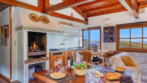 A spacious living area with a fireplace, wooden beams, a dining table set with plates, glasses, and a bowl of salad. - 5-bed Villa, Two Pools, Puerto Pollenca (Pollença)