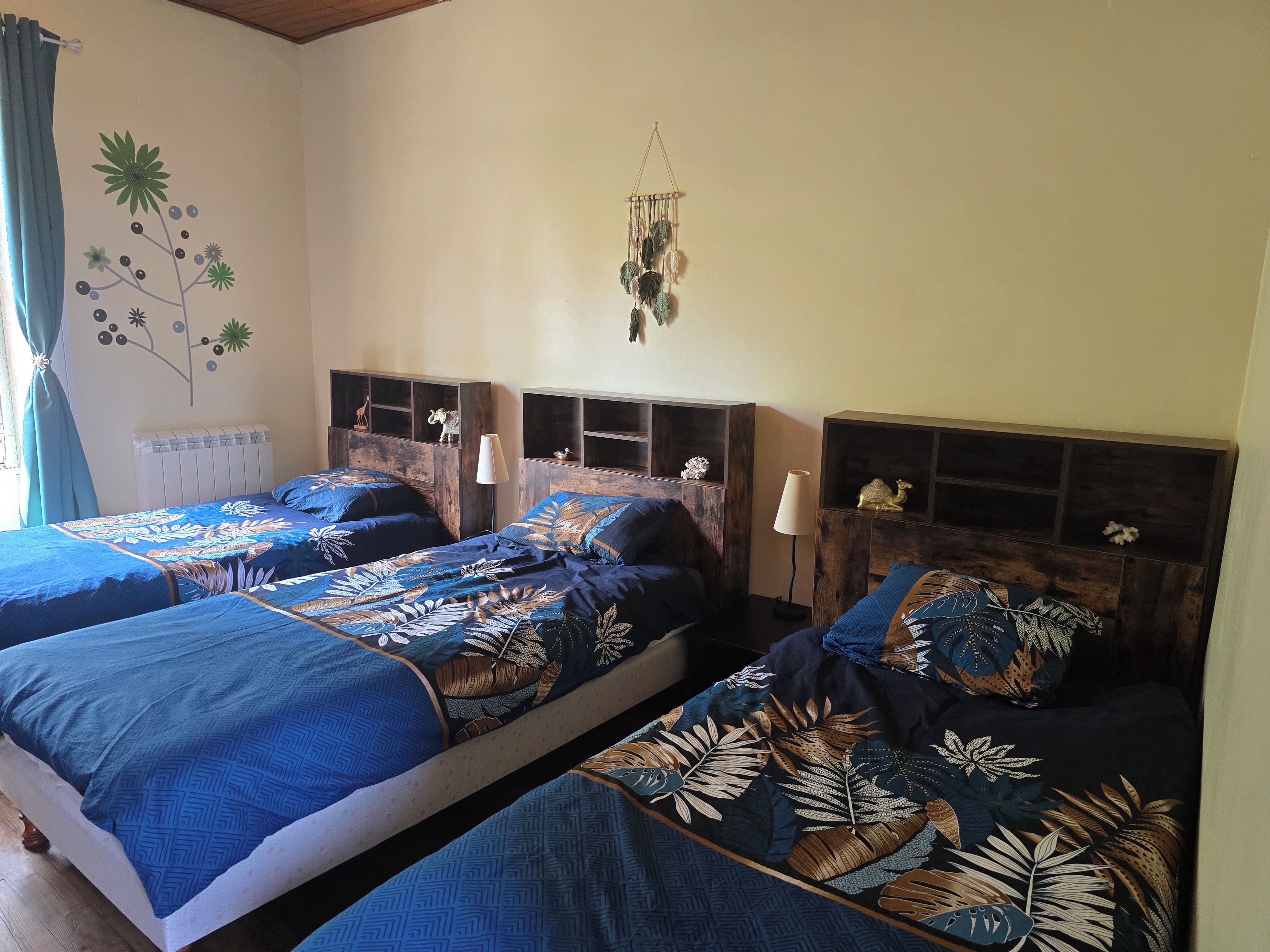 Une chambre avec deux lits, chacun recouvert d’une couvre-lit à motifs, une étagère fixée au mur avec des objets décoratifs, et une décoration en macramé suspendue.