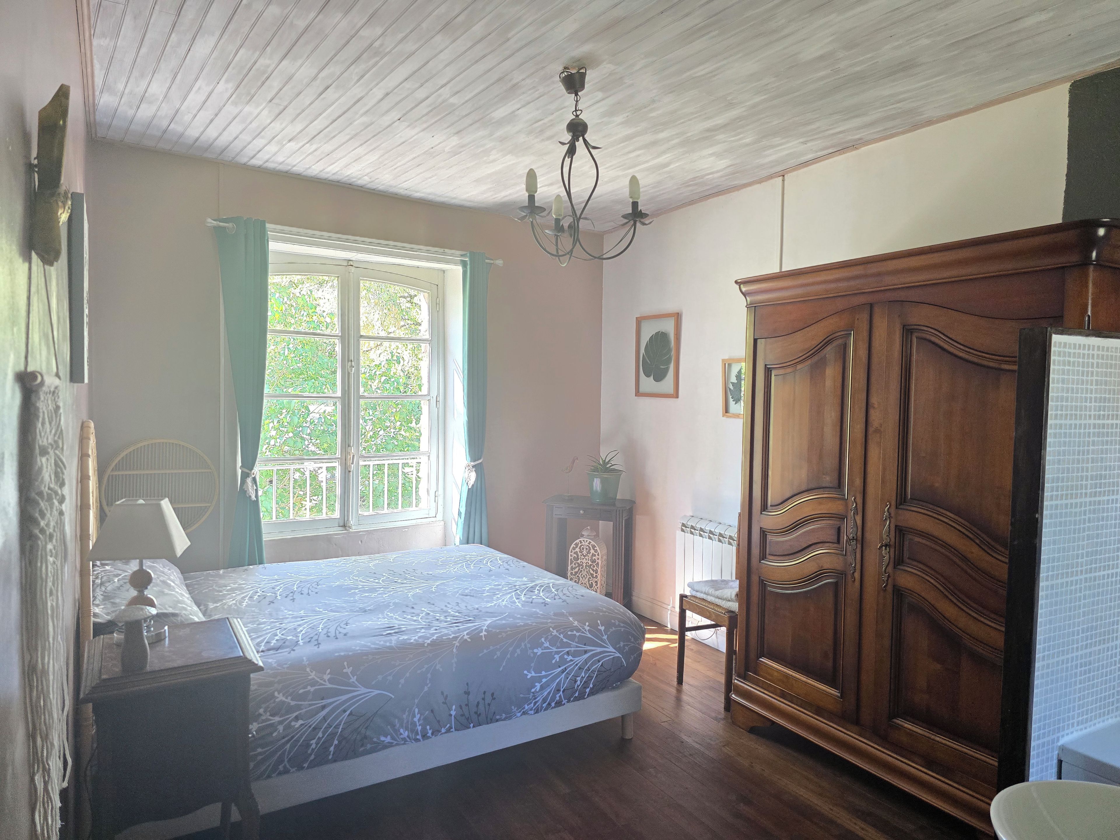 Un dormitorio con cama, armario de madera, ventana con cortinas, radiador y candelabro.