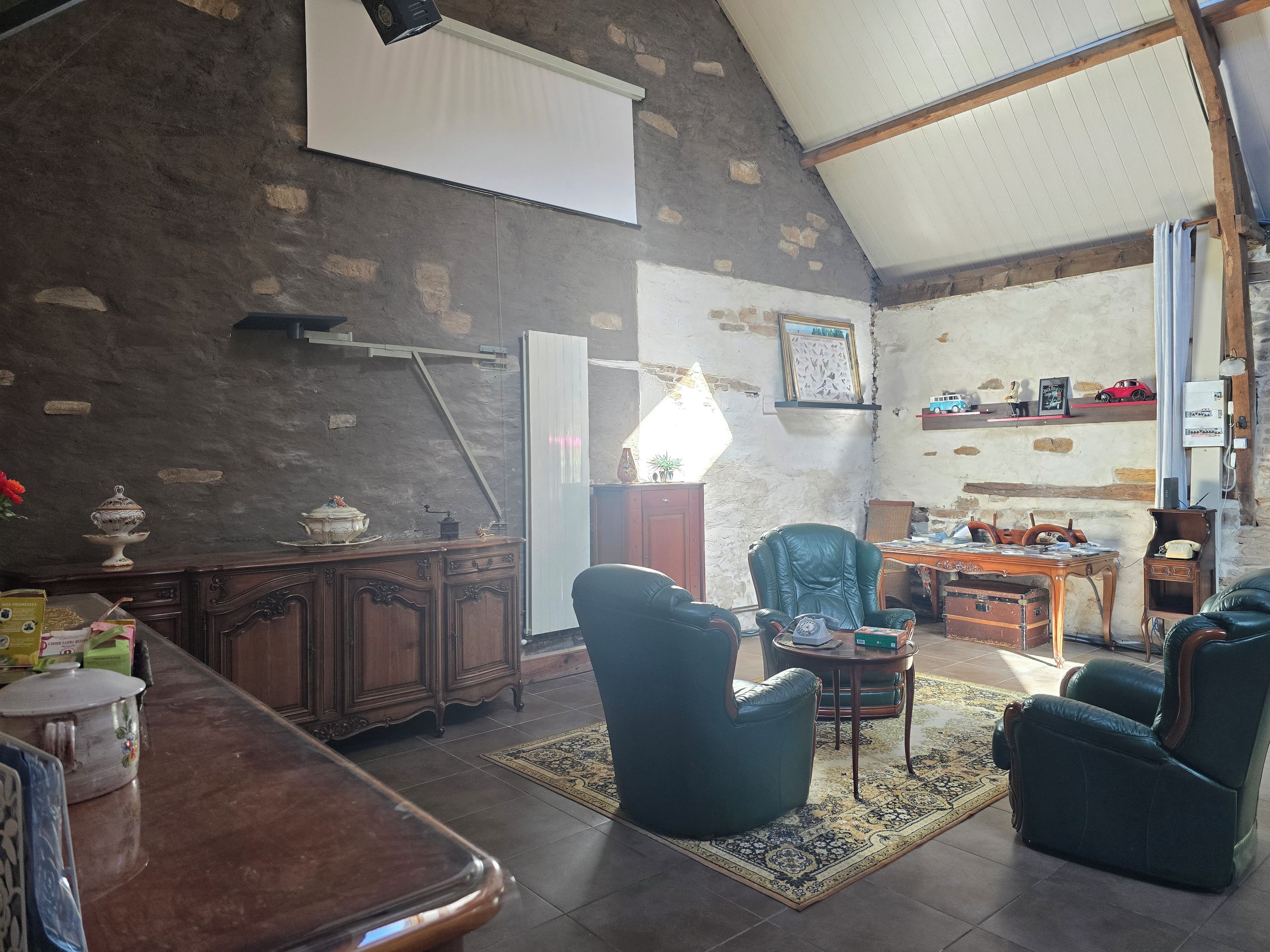 Une pièce spacieuse avec un plafond haut, une grande fenêtre et un mur en pierre rustique. On y trouve un bureau en bois avec une lampe, un fauteuil vert et une petite table avec une chaise.