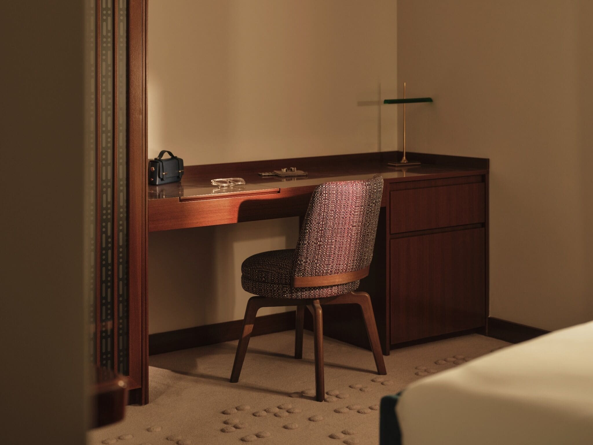 A hotel room with a wooden desk, a patterned chair, a small table, and a lamp.