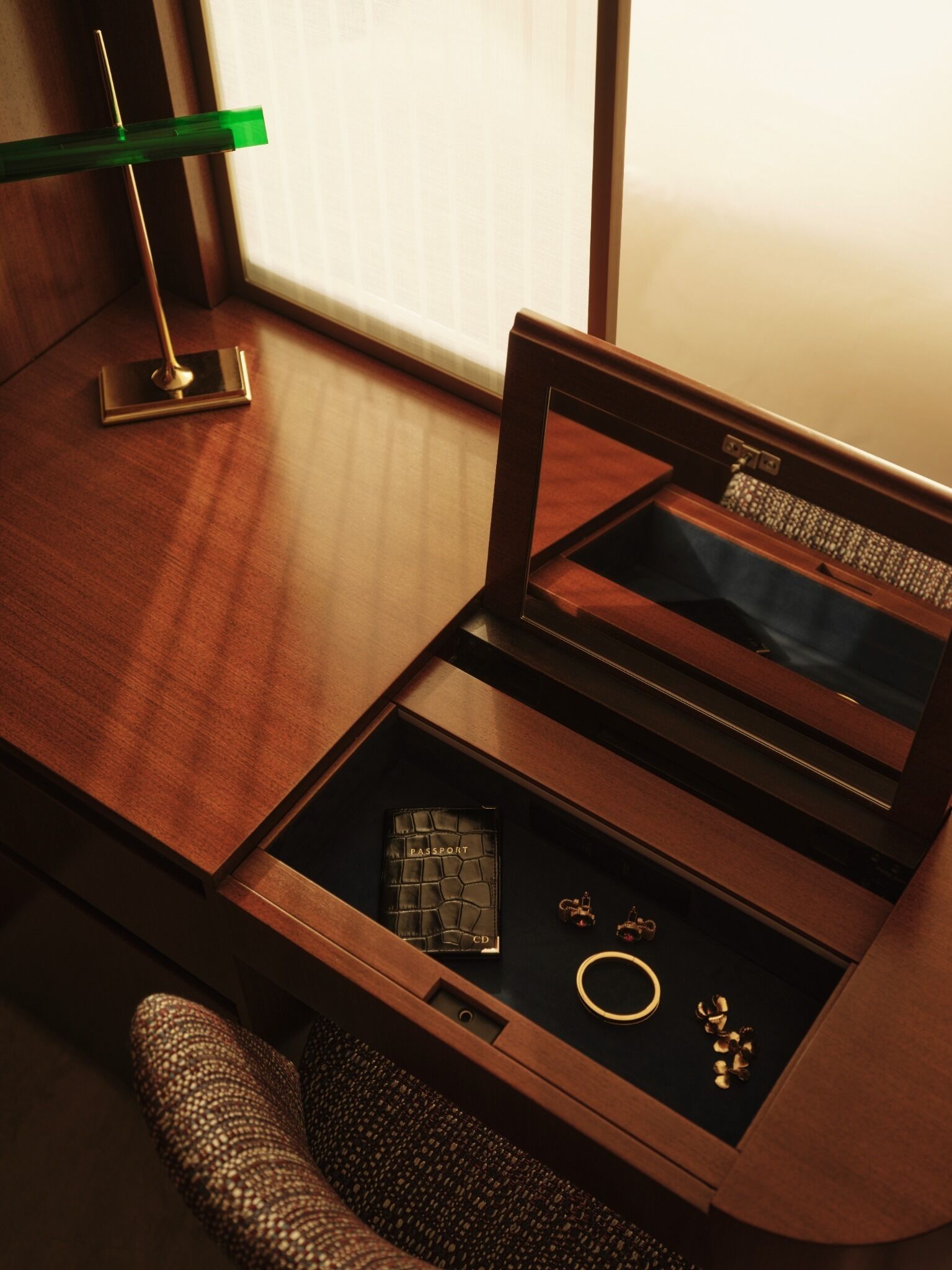 Un escritorio de madera con una lámpara de escritorio verde, un pasaporte con cubierta de cuero negro, un CD, un anillo de oro y un par de pendientes.
