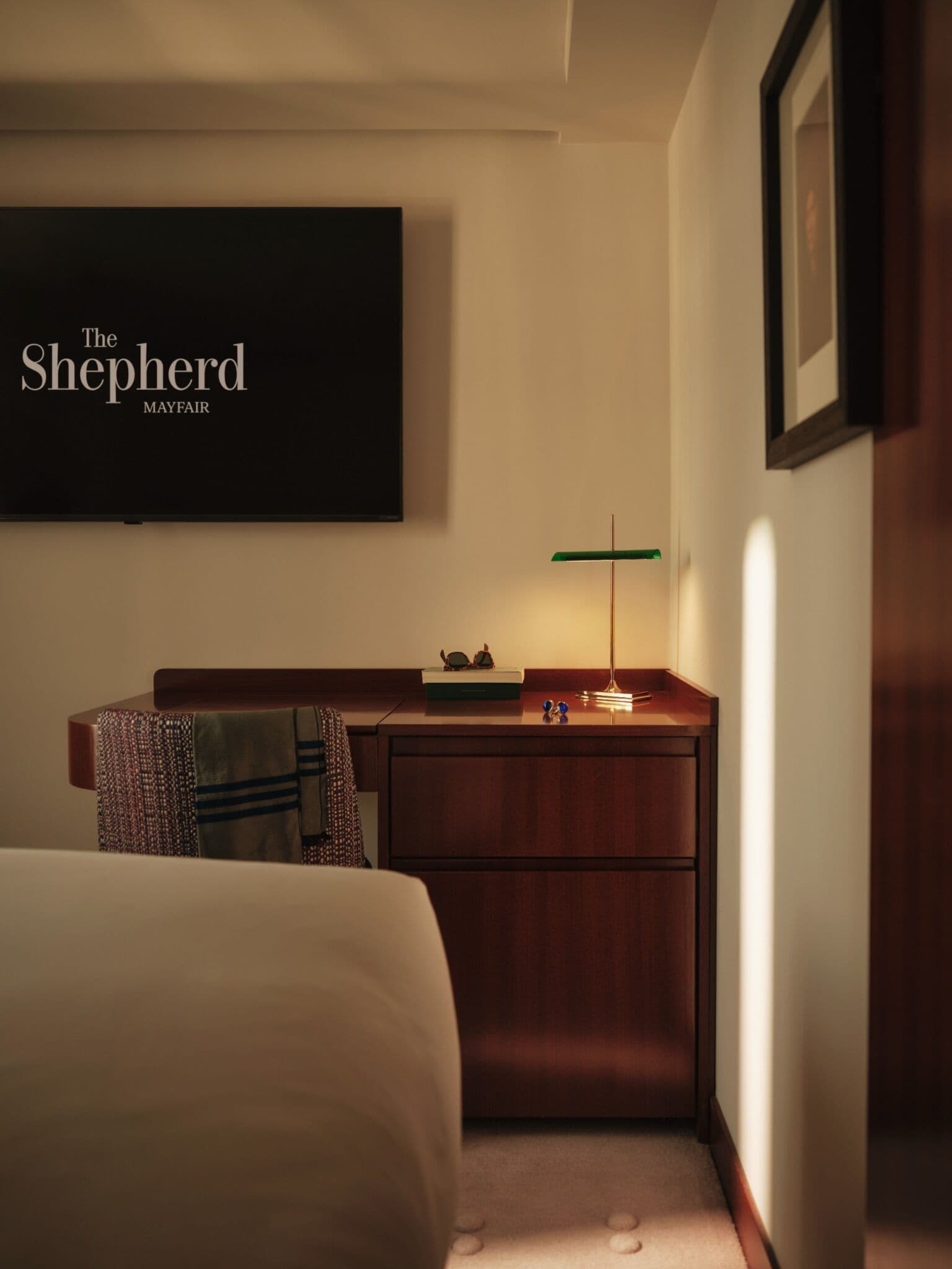 A hotel room with a bed, a desk with a lamp, a framed picture on the wall, and a sign reading 'The Shepherd Mayfair'.