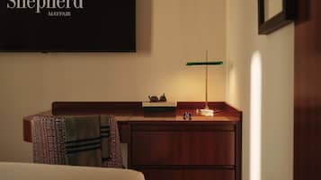 A hotel room with a bed, a desk with a lamp, a framed picture on the wall, and a sign reading 'The Shepherd Mayfair'.