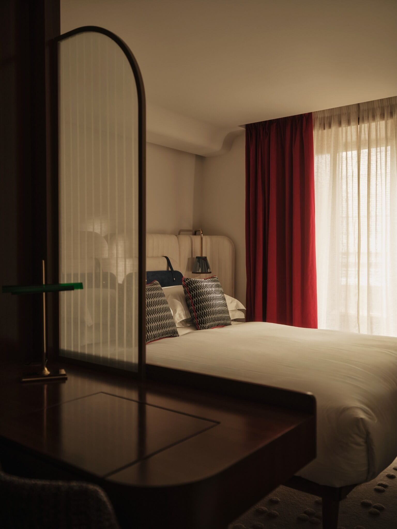 A hotel room with a bed, a bedside table, a lamp, and a window with curtains.