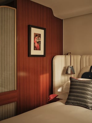 A hotel room with a wooden panel wall, a framed picture, a bedside lamp, a patterned pillow, and a red object on the bed. - The Shepherd Mayfair (London)